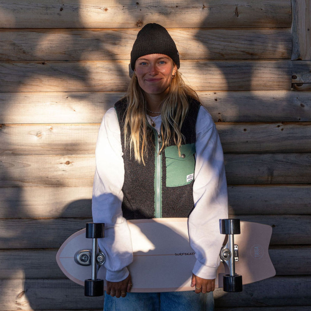 Person wearing a fisherman taiga beanie holding a skateboard against a wooden wall