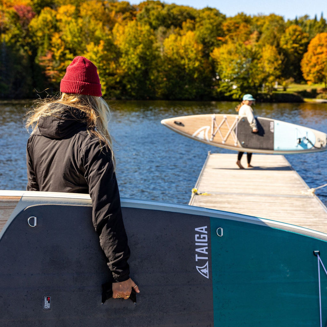 Person looking at another person with a paddleboard on a dock by a lake with autumn trees.