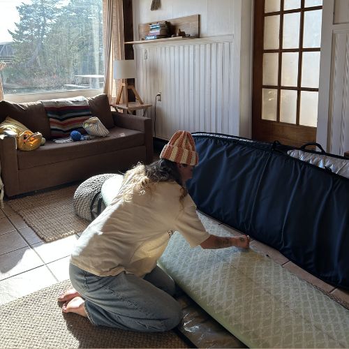 Person waxing a surfboard in a living room with a couch and window in the background.