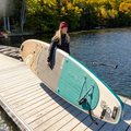 Woman with a teal and white paddleboard on a dock by a body of water.