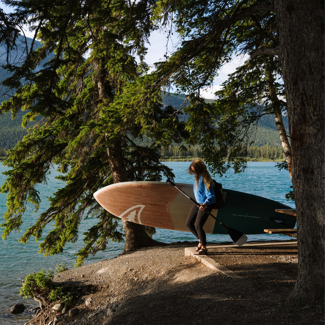 Person holding a paddle board by a lake with trees and mountains in the background