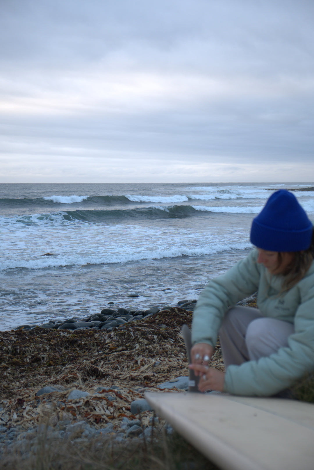 Person on a beach with a surfboard and blue beanie, ocean waves in the background