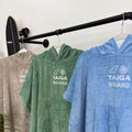 Three poncho towels in gray, green, and blue hanging on a rack with a skateboard, all featuring the 'TAIGA BOARD' logo.