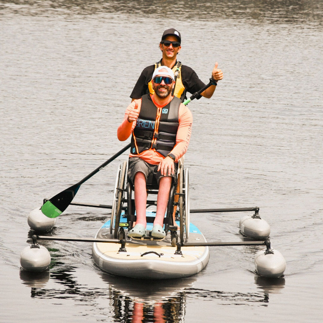 Two people on a paddleboard with one in a wheelchair, on a calm body of water.