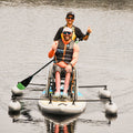 Two people on a paddleboard with one in a wheelchair, on a calm body of water.