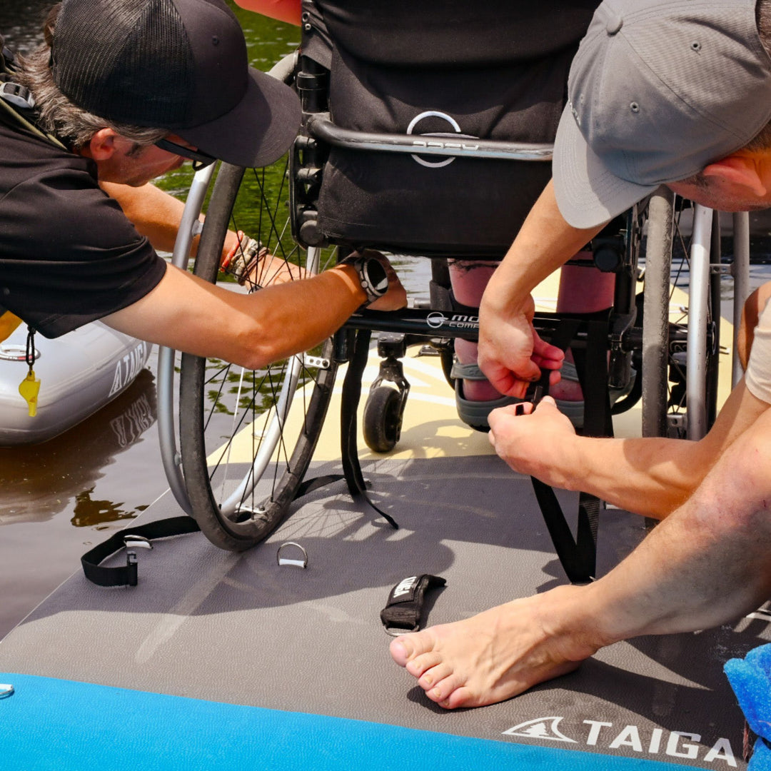 Two individuals assisting a person in a wheelchair by a body of water, with 'TAIGA' branding visible.