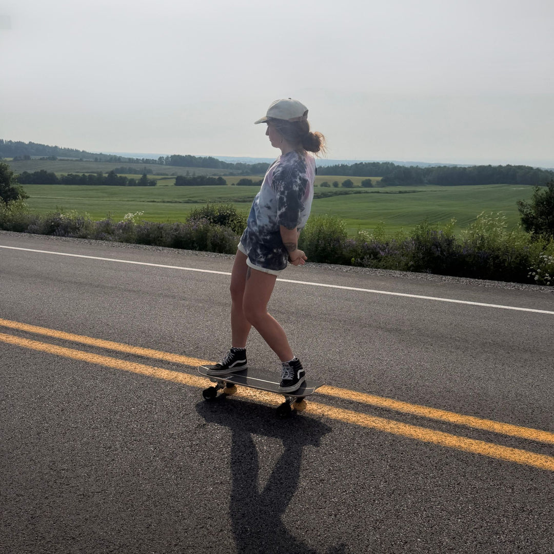 A person stands on a skateboard on a paved road with a green field and trees in the background, wearing a gray patterned t-shirt, white shorts, and black sneakers.