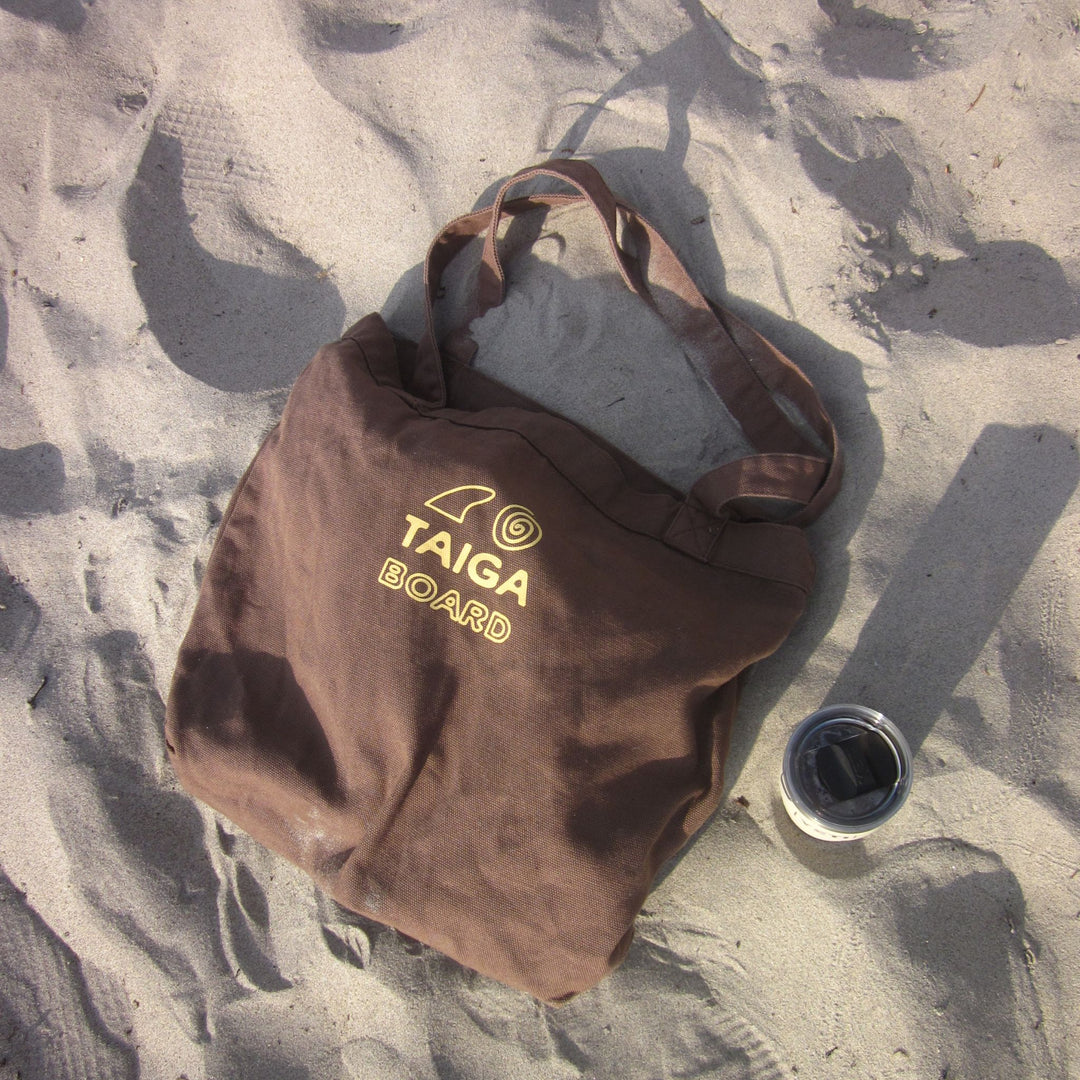 Brown tote bag with 'TAIGA BOARD' logo on sandy ground